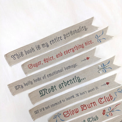 Hand painted linen ribbon bookmarks that read "Sugar, spice, and everything nice" "Slow Burn Club" "Enemies to Lovers Club" and more phrases