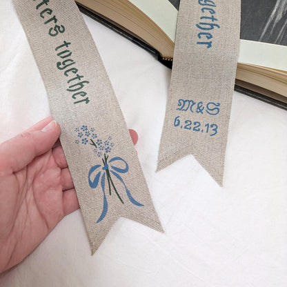 Hand painted linen ribbon bookmark that reads "To many more chapters together" with either initials and wedding anniversary date motif or forget-me-not-flower motif