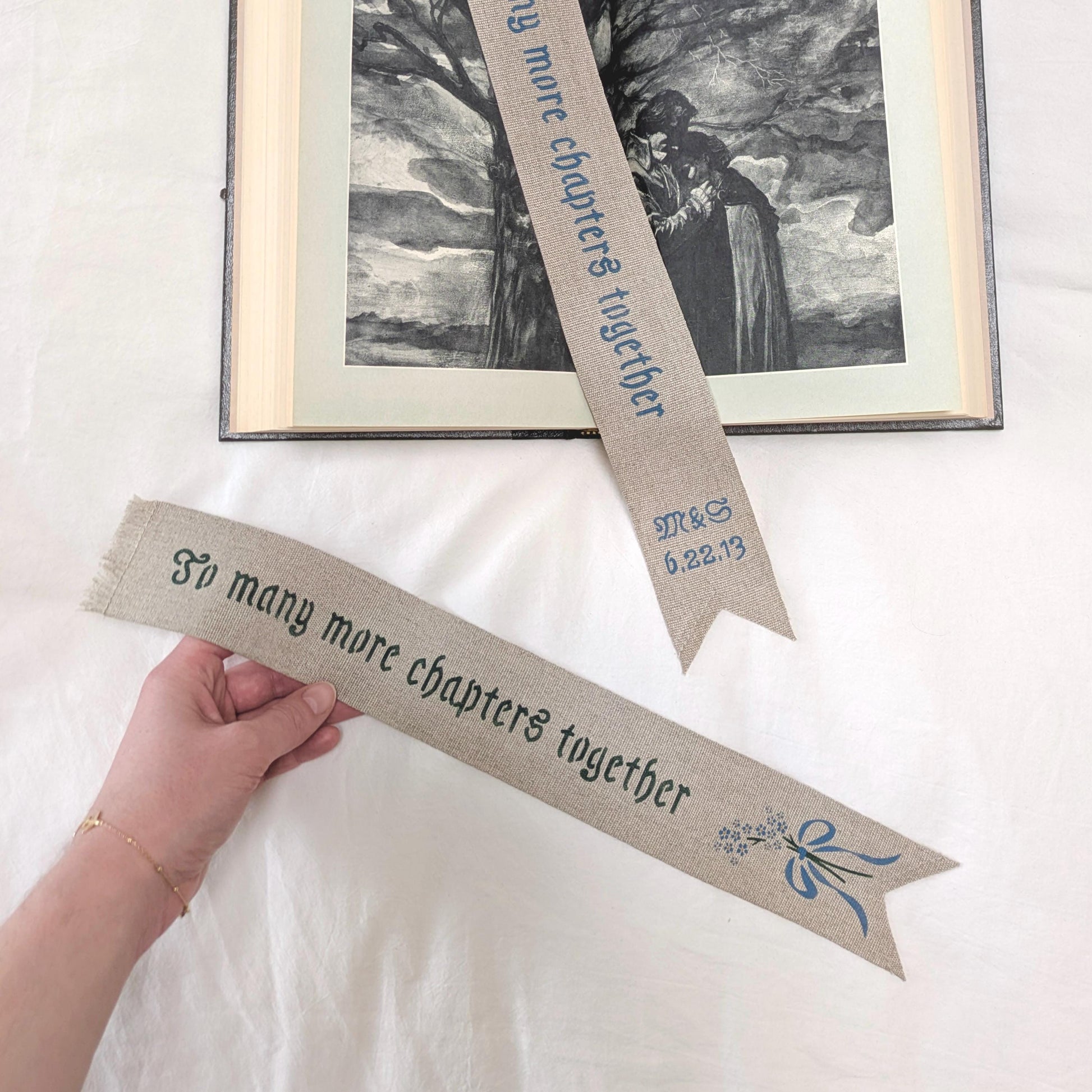 Hand painted linen ribbon bookmark that reads "To many more chapters together" with either initials and wedding anniversary date motif or forget-me-not-flower motif